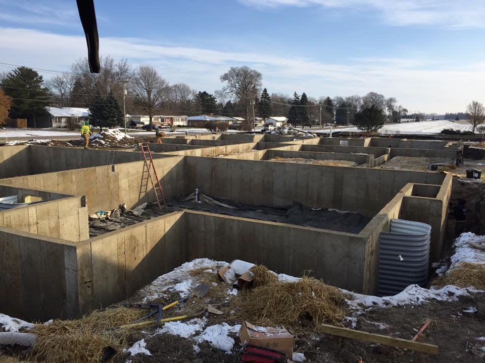 Overview of multiple new residential poured concrete foundations under construction in winter conditions with snow and straw, with workers in the distance.