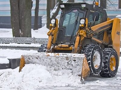 Yellow skid steer loader equipped with a snow bucket attachment, actively plowing and removing snow from a sidewalk or paved area.