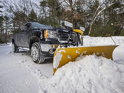 Heavy-duty black pickup truck with a bright yellow snowplow attachment clearing a snow-covered driveway or road in winter.